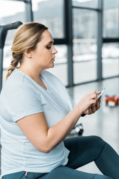 Overweight girl using smartphone while sitting in gym — Stock Photo