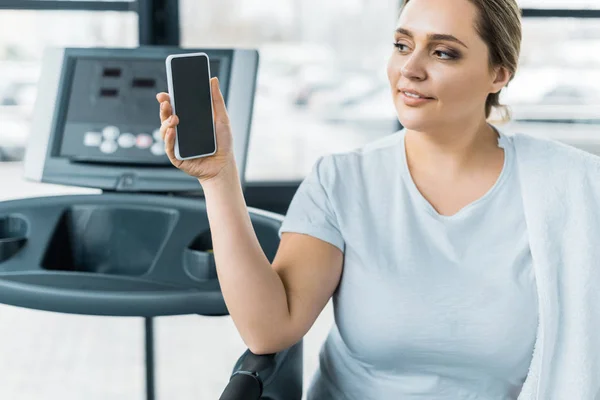 Alegre chica con sobrepeso mirando el teléfono inteligente con pantalla en blanco en el gimnasio - foto de stock