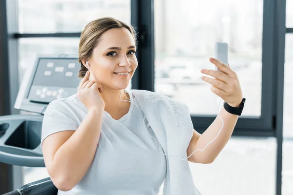 Smiling overweight girl holding smartphone while listening music in earphones — Stock Photo