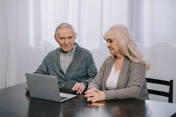 Bouleversé couple âgé assis à la table et en utilisant un ordinateur portable à la maison — Photo de stock
