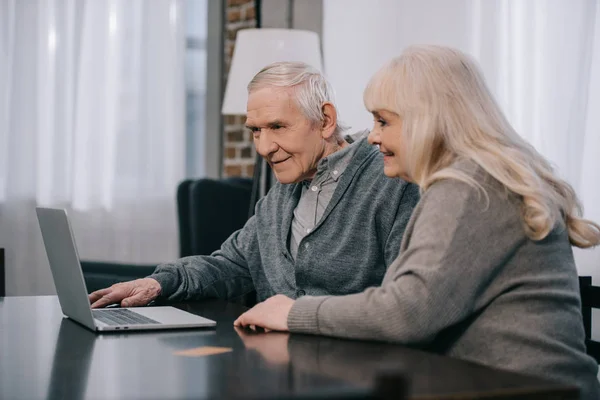 Heureux couple de personnes âgées assis à la table et en utilisant un ordinateur portable à la maison — Photo de stock