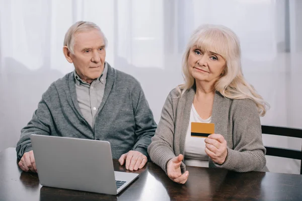 Seniorenpaar sitzt am Tisch und hält Kreditkarte in der Hand, während es zu Hause Laptop benutzt — Stockfoto