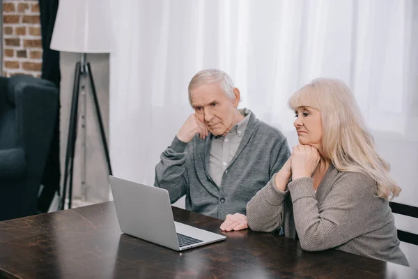 Senior couple sitting at table, propping chins with hands and using laptop at home — Stock Photo