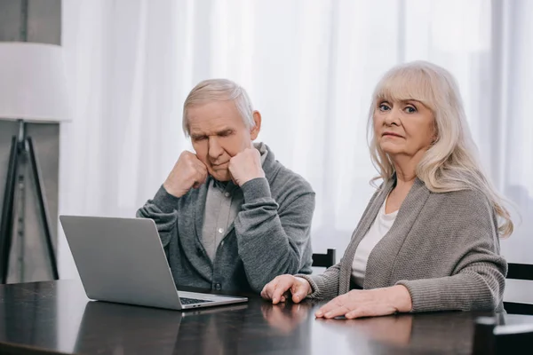 Upset senior couple with grey hair sitting at table and using laptop at home — Stock Photo