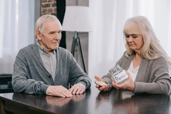 Upset senior couple sitting at table and holding empty glass jar with 'pension' lettering — Stock Photo