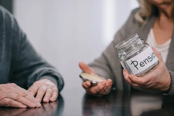 Cropped view of senior couple sitting at table and holding empty glass jar with 'pension' lettering — Stock Photo