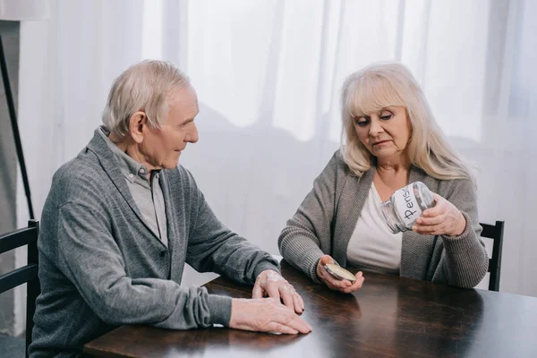 Sad senior couple sitting at table and holding empty glass jar with 'pension' lettering — Stock Photo