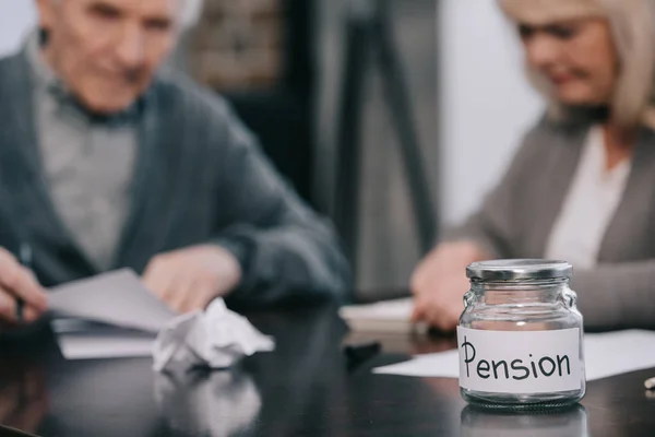 Selective focus of empty glass jar with 'pension' lettering on table with senior couple on background — Stock Photo