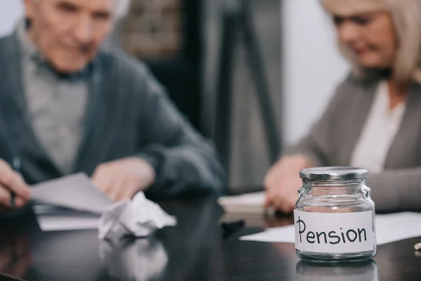 Enfoque selectivo de frasco de vidrio vacío con letras de 'pensión' en la mesa con pareja mayor en el fondo - foto de stock