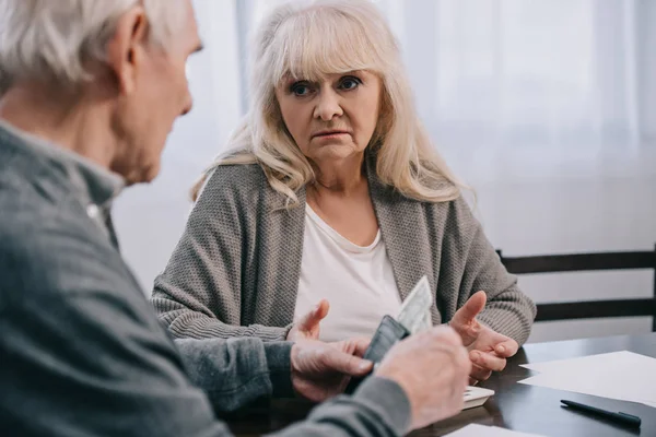 Pareja de ancianos disgustados en ropa casual sosteniendo el dinero y sentado en la mesa - foto de stock