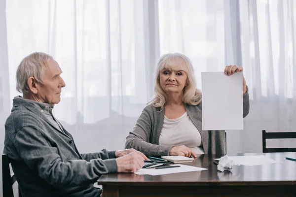 Senior woman holding empty paper and looking at camera while sitting at table with man — Stock Photo