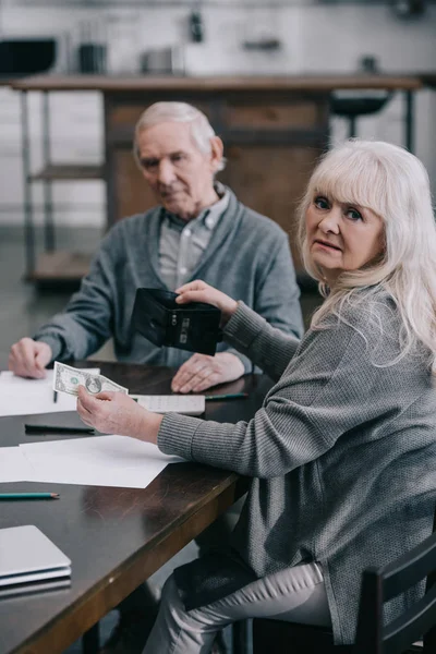 Seniorin hält Geldbörse und Geld in der Hand und blickt in Kamera, während sie mit Mann am Tisch sitzt — Stockfoto