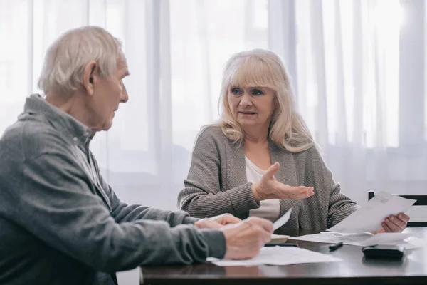 Senior couple in casual clothes sitting at table and holding bills — Stock Photo