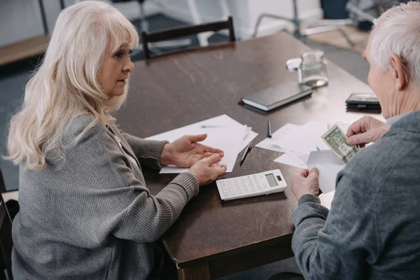 Casal sênior sentado à mesa com calculadora, dinheiro e papelada — Fotografia de Stock