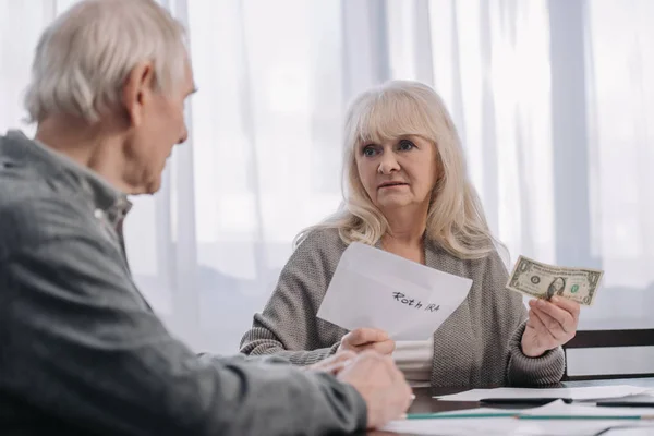 Senior couple holding envelope with 'roth ira' lettering and dollar banknote at home — Stock Photo