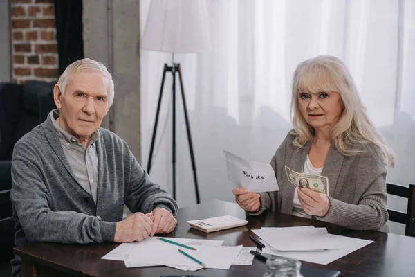 Senior couple holding money, envelope with 'roth ira' lettering and looking at camera — Stock Photo