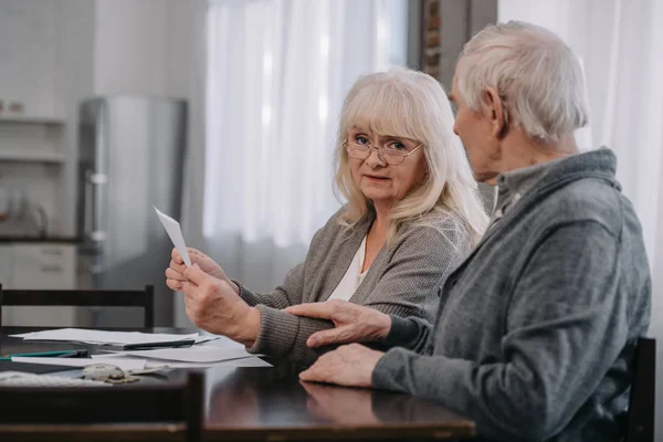Senior femme en lunettes tenant des factures et regardant la caméra tout en étant assis à la table avec l'homme — Photo de stock