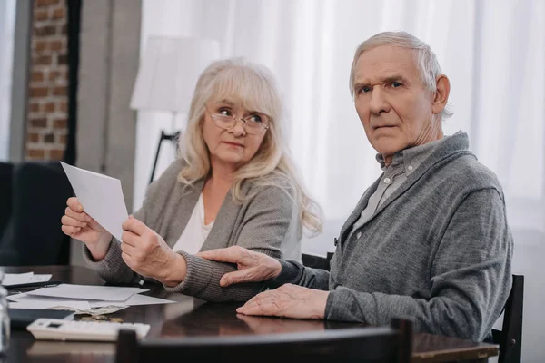 Couple aîné en vêtements décontractés tenant des factures tout en étant assis à table — Photo de stock