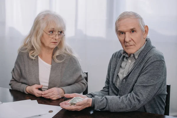 Couple aîné en vêtements décontractés assis à table et tenant de l'argent dans les mains — Photo de stock
