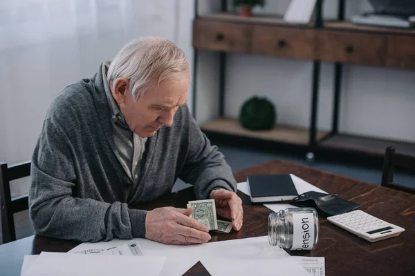 Senior man sitting at table with money, paperwork and glass jar with 'pension' lettering — Stock Photo