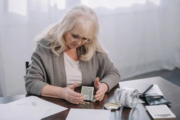 Mujer mayor sentada en la mesa con dinero, papeleo y frasco de vidrio vacío con palabra 'pensión' - foto de stock