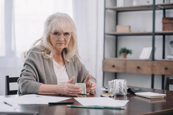 Femme âgée assise à table avec de l'argent, de la paperasse et un bocal en verre vide avec mot 