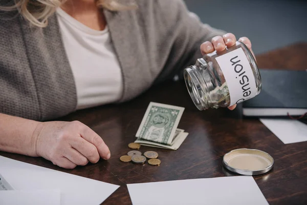 Cropped view of senior woman holding glass jar with 'pension' word while counting money at home — Stock Photo
