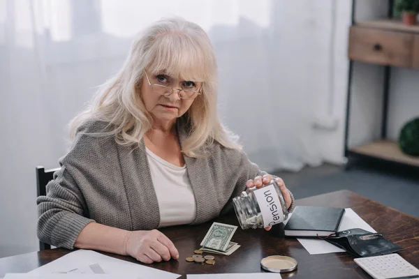 Senior woman looking at camera, holding glass jar with 'pension' word and counting money — Stock Photo