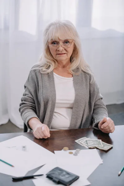 Senior woman in glasses sitting at table and looking at camera while counting money at home — Stock Photo