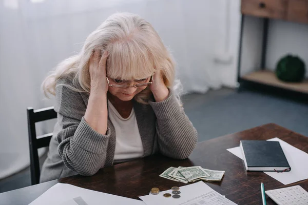 Втомлена старша жінка з руками на голові має головний біль під час підрахунку грошей вдома — стокове фото