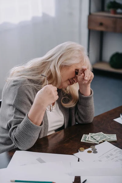 Cansada mujer mayor sentada en la mesa y con dolor de cabeza mientras cuenta dinero en casa - foto de stock