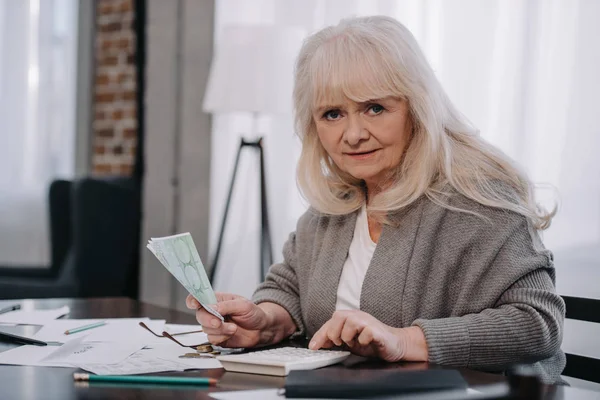 Mujer mayor sentada en la mesa, usando calculadora y contando dinero - foto de stock