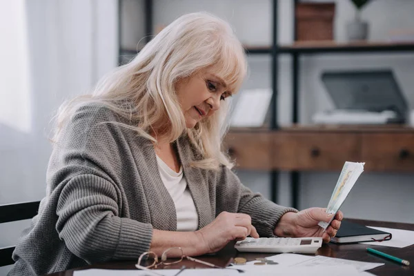 Seniorin sitzt am Tisch, benutzt Taschenrechner und zählt Geld — Stockfoto