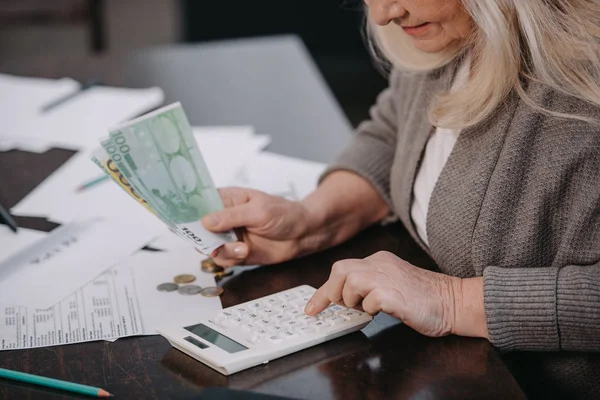 Обрізаний вид на старшу жінку, що сидить за столом, використовуючи калькулятор і рахуючи гроші — стокове фото