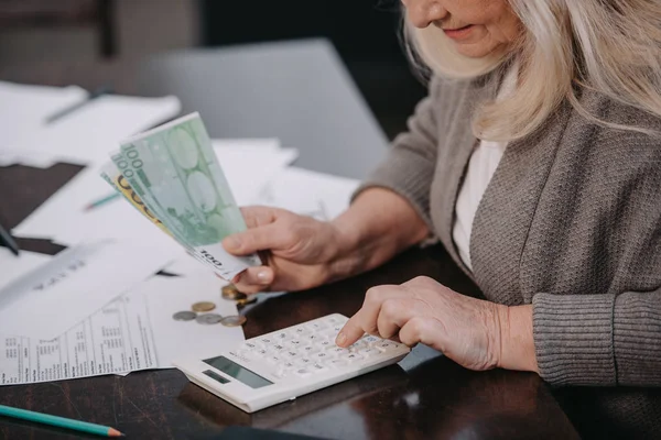 Vista recortada de la mujer mayor sentada en la mesa, usando calculadora y contando dinero - foto de stock