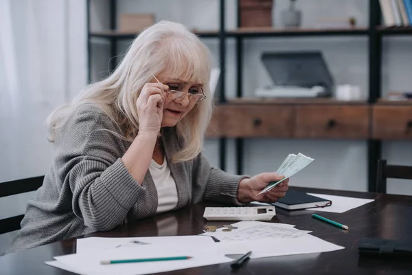 Seniorin sitzt mit Papierkram am Tisch, hält Brille und zählt Geld — Stockfoto