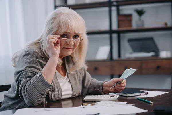 Mujer mayor sentada en la mesa con papeleo, mirando a la cámara y contando dinero - foto de stock