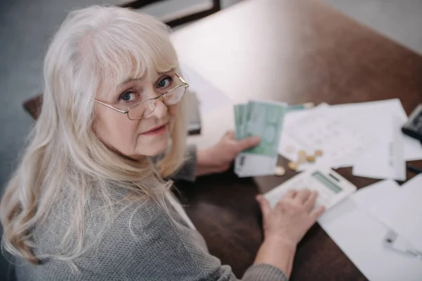 Seniorin sitzt am Tisch, blickt in Kamera, benutzt Taschenrechner und zählt Geld — Stockfoto