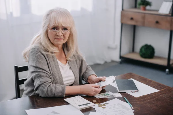 Seniorin sitzt mit Papierkram am Tisch, blickt in Kamera und zählt Geld — Stockfoto