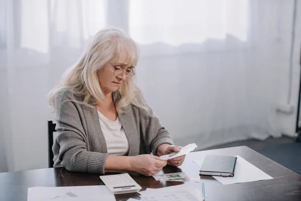 Старшая женщина сидит за столом с бумагами и подсчитывает деньги — стоковое фото