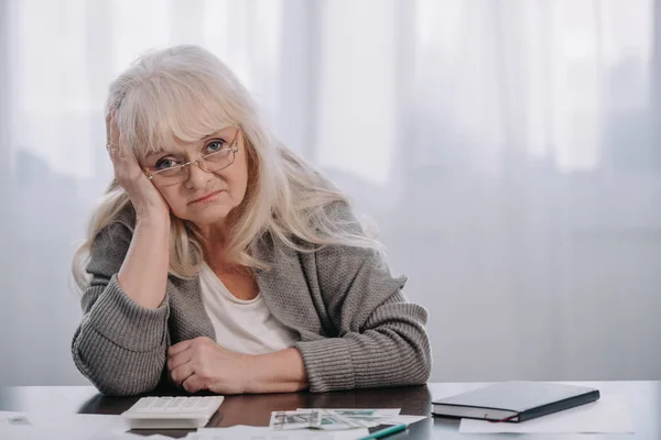 Femme âgée assise à table avec de l'argent, regardant la caméra et ayant mal à la tête — Photo de stock