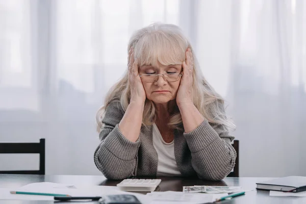 Gestresste Seniorin sitzt mit Geld und Papieren am Tisch und hat zu Hause Kopfschmerzen — Stockfoto