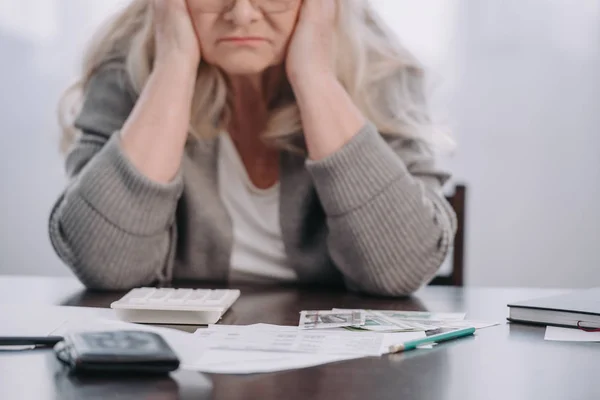 Abgeschnittene Ansicht einer Seniorin, die mit Geld und Papieren am Tisch sitzt, während sie zu Hause Kopfschmerzen hat — Stockfoto