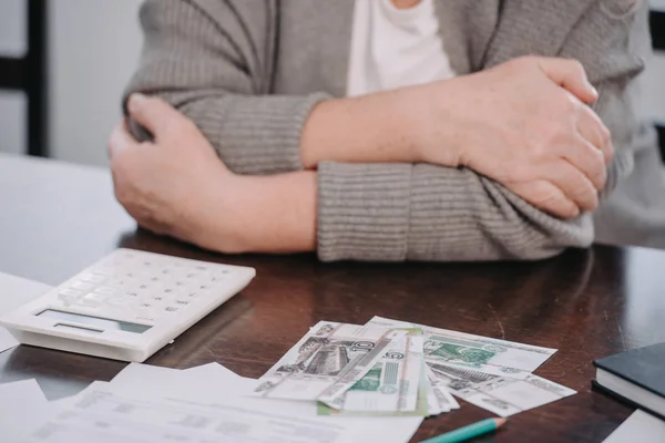 Обрізаний вид на старшу жінку з перехрещеними руками сидить за столом з грошима і калькулятором — стокове фото