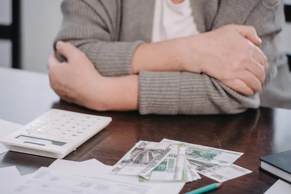 Vue recadrée de la femme âgée avec les bras croisés assis à la table avec de l'argent et calculatrice — Photo de stock