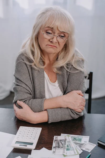 Donna anziana sconvolta con le braccia incrociate seduta a tavola con soldi e calcolatrice — Foto stock