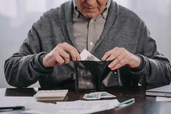 Visão cortada do pensionista masculino sentado à mesa com papelada e colocando dinheiro na carteira — Fotografia de Stock