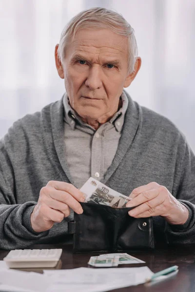 Male pensioner sitting at table, looking at camera and putting money in wallet — Stock Photo