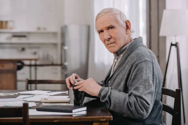 Pensionné mâle assis à table avec de la paperasse, regardant la caméra et mettant de l'argent dans le portefeuille — Photo de stock