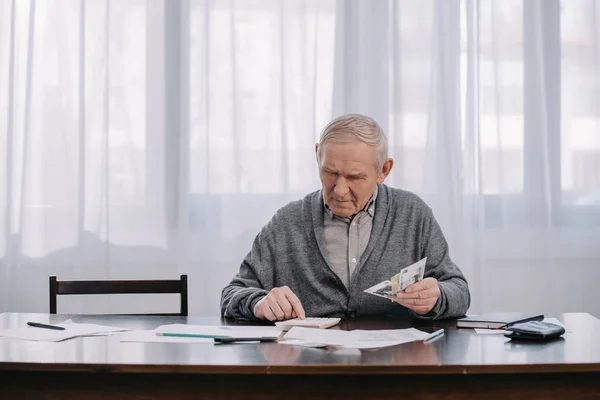 Uomo anziano seduto a tavola con scartoffie e utilizzando la calcolatrice mentre il conteggio dei soldi — Foto stock